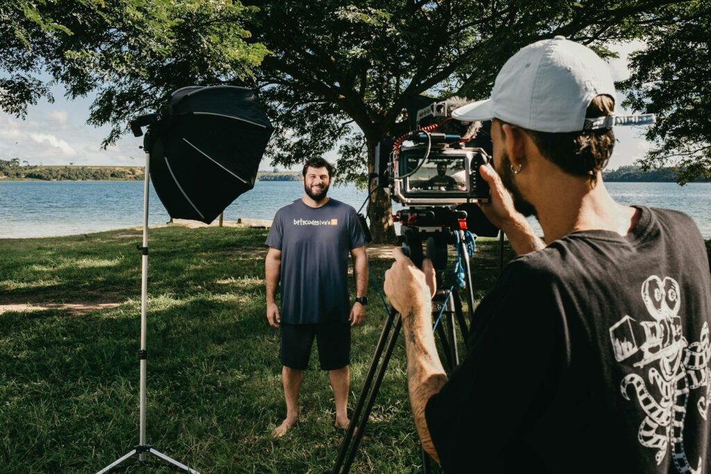 Filmmaker with video camera on tripod shooting video of smiling man standing on glade on coast of pond