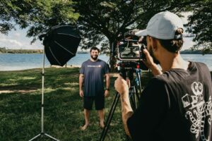 Filmmaker with video camera on tripod shooting video of smiling man standing on glade on coast of pond