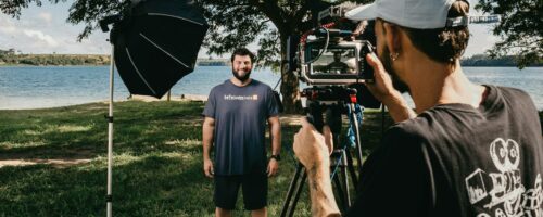 Filmmaker with video camera on tripod shooting video of smiling man standing on glade on coast of pond