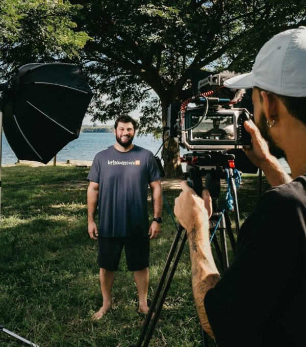 Filmmaker with video camera on tripod shooting video of smiling man standing on glade on coast of pond