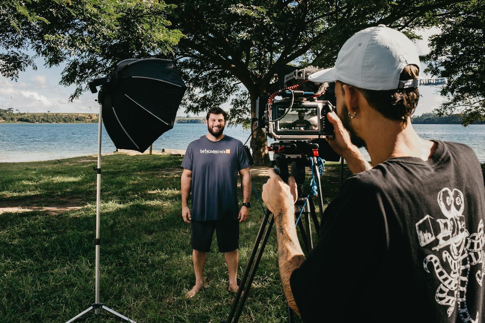 Filmmaker with video camera on tripod shooting video of smiling man standing on glade on coast of pond